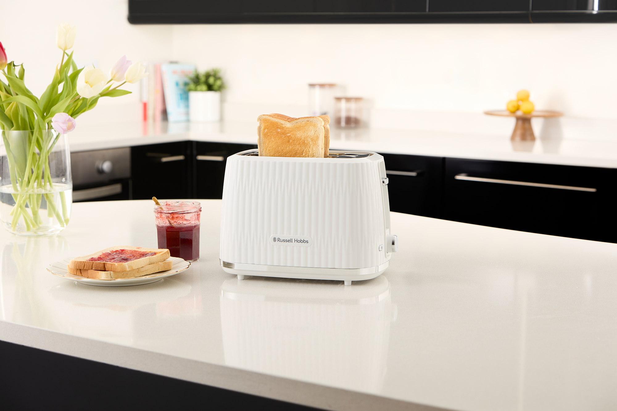 Fehér Russell Hobbs kenyérpirító kenyérrel, pulton. Üvegek és virágok láthatók.