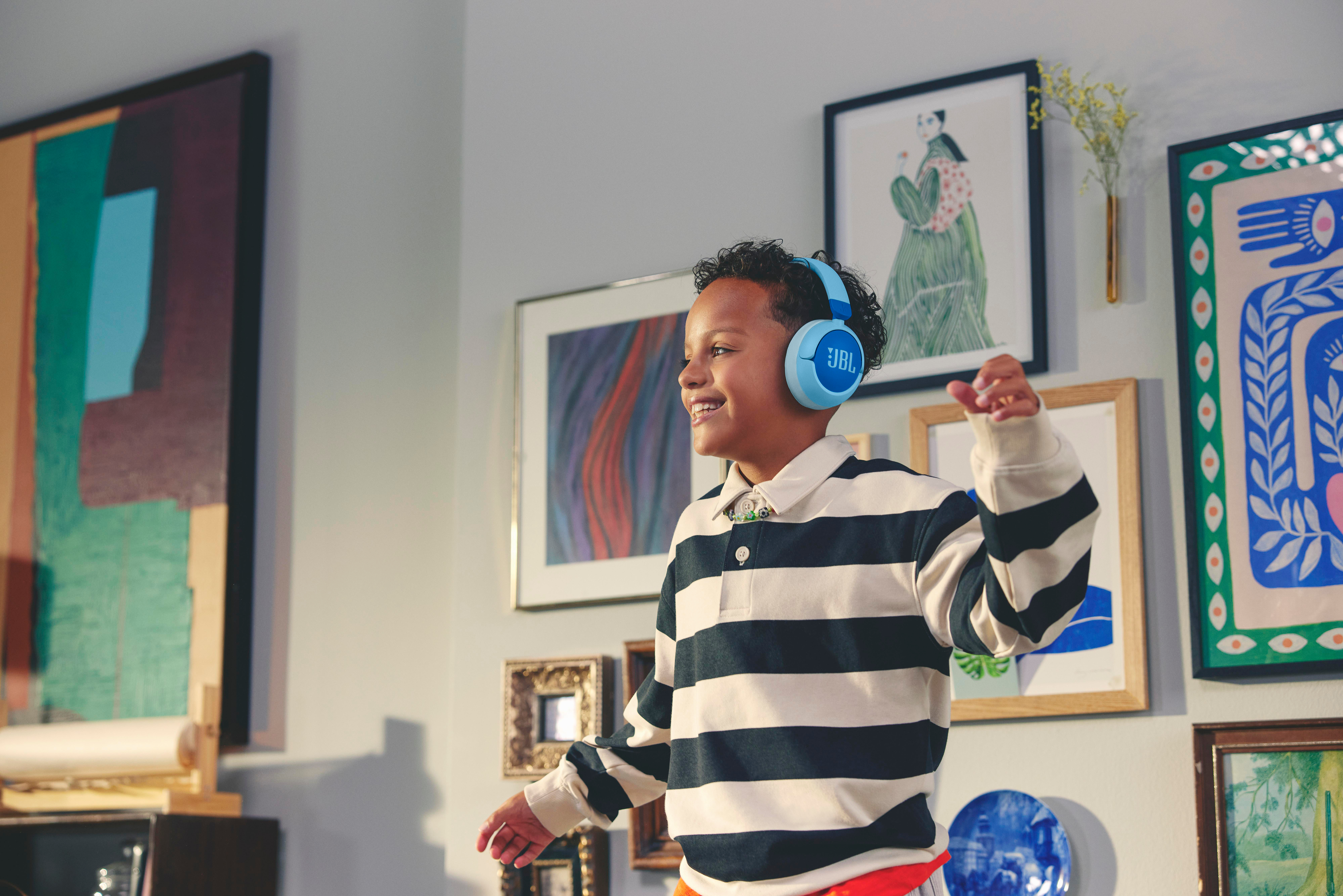 Junge trägt blaue JBL-Kopfhörer, lächelt und tanzt in einem Raum mit Kunst.