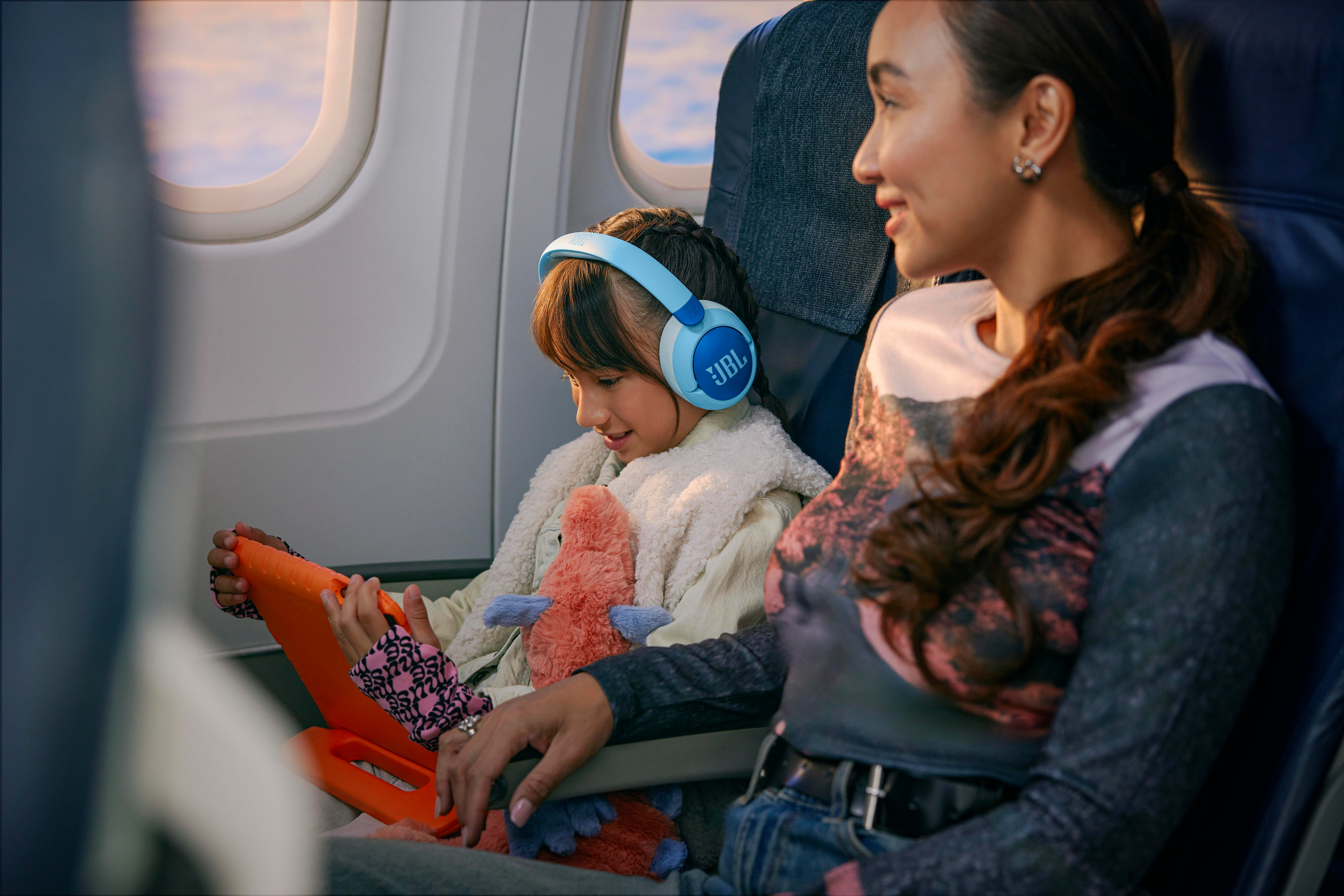 Mädchen im Flugzeug mit blauen Kopfhörern, schaut auf Tablet. Frau neben ihr.