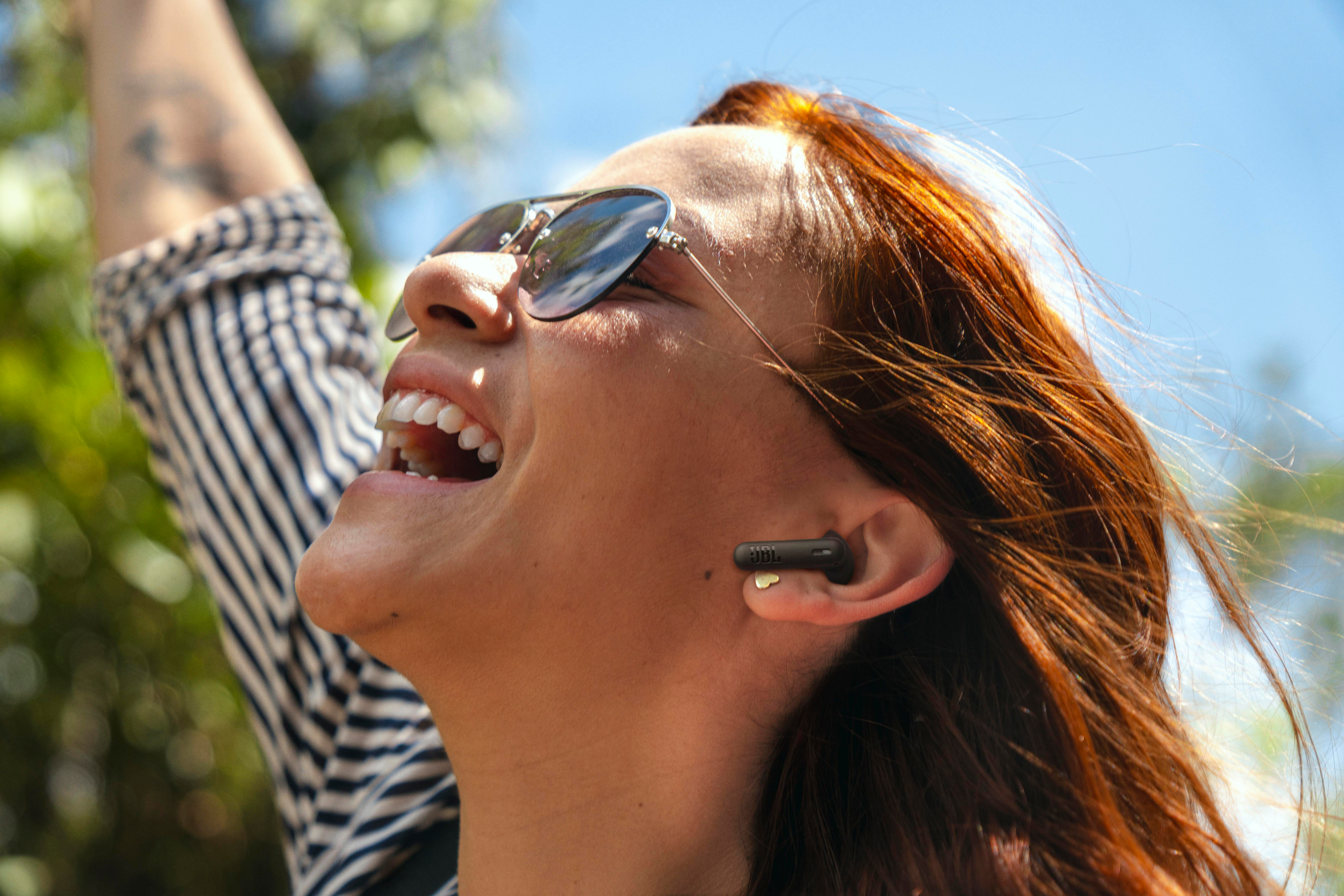 Frau mit Sonnenbrille lächelt, Ohrhörer im Ohr, greift unter blauem Himmel.