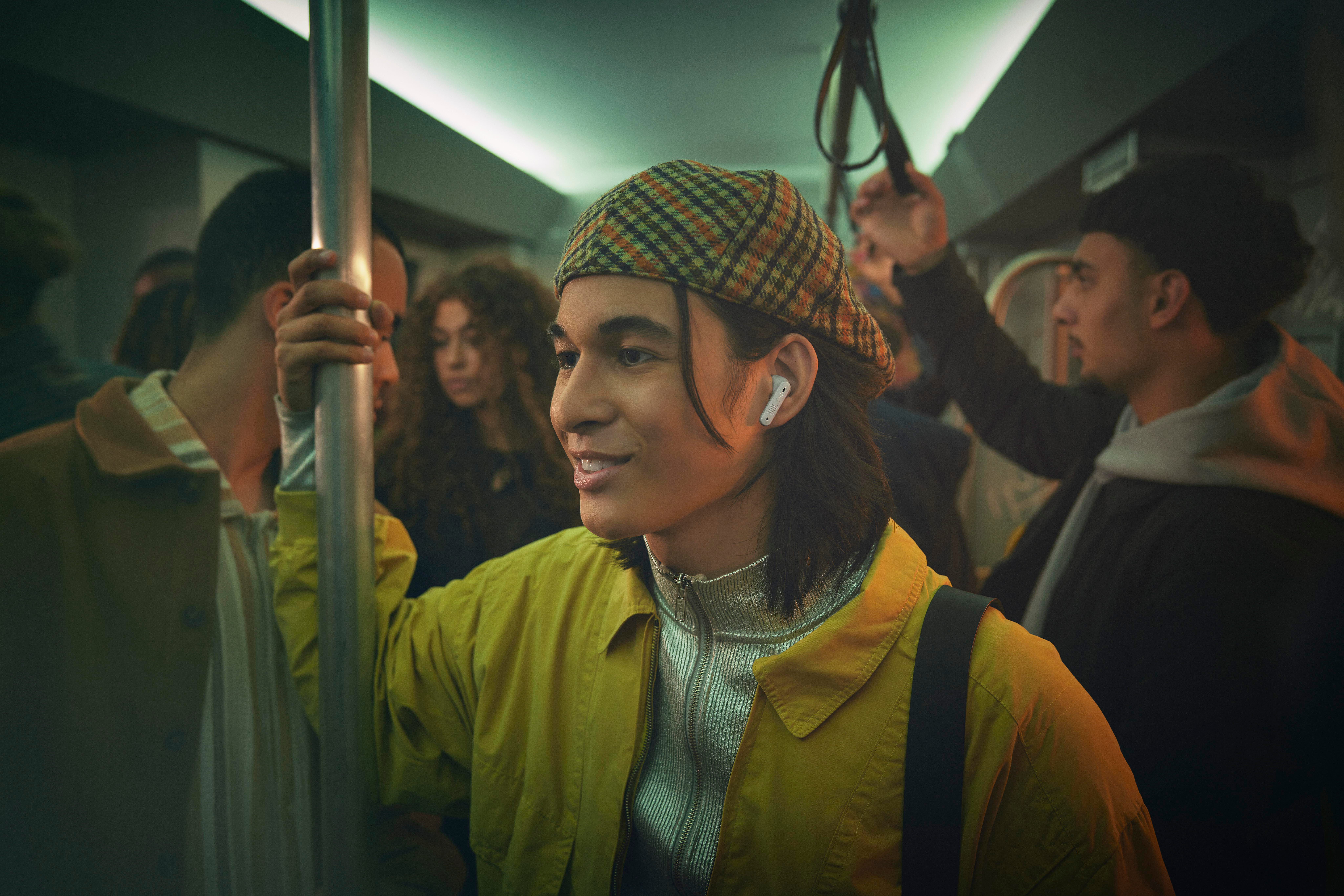 Ein Mann mit Ohrhörern in der U-Bahn, trägt eine gelbe Jacke und Hut.