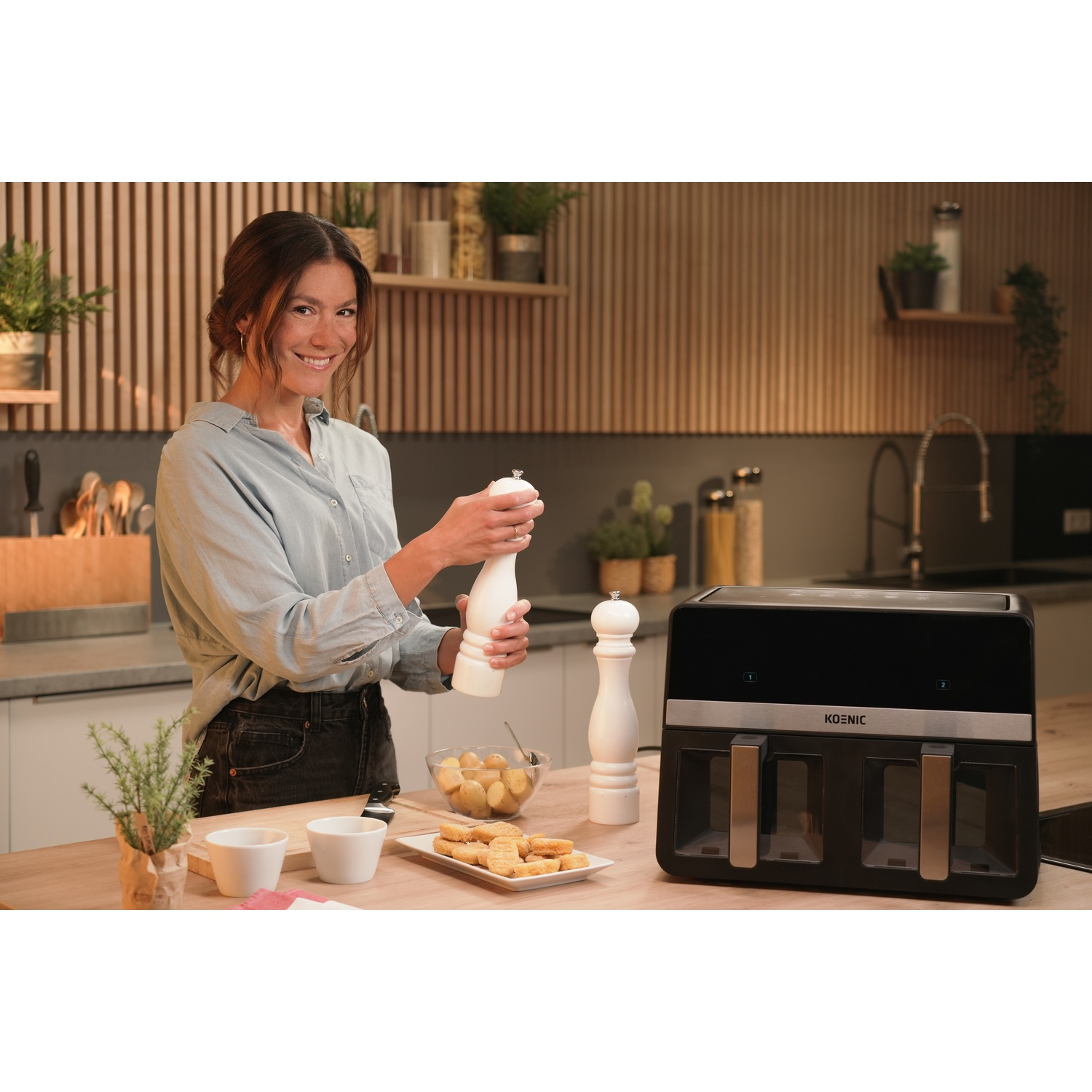 Donna in cucina con macinapepe. La cucina ha mobili in legno, piani di lavoro bianchi e una friggitrice ad aria.
