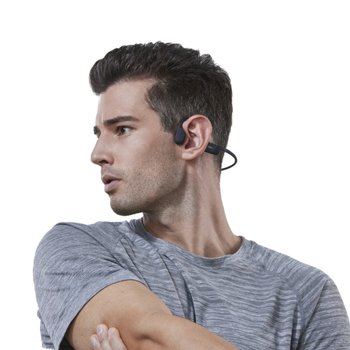 Un hombre con auriculares de conducción ósea, mirando hacia otro lado. Lleva una camisa gris.