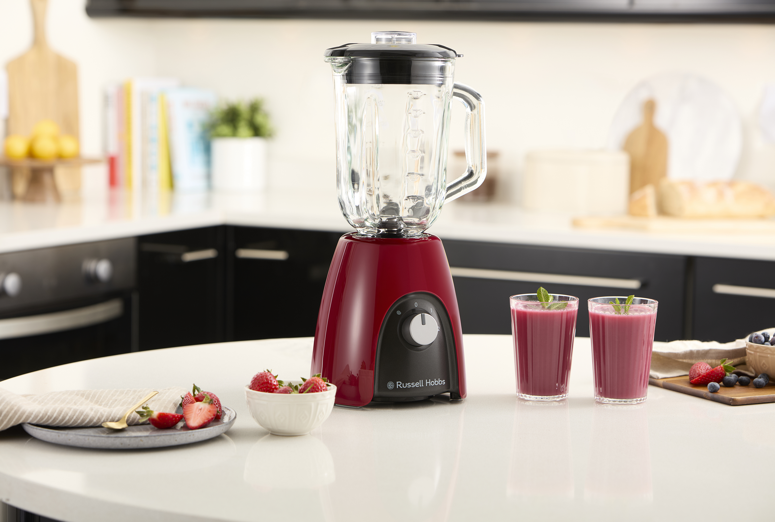 Piros turmixgép üvegkancsóval, eperrel és két pohár lila smoothie-val a fehér pulton.