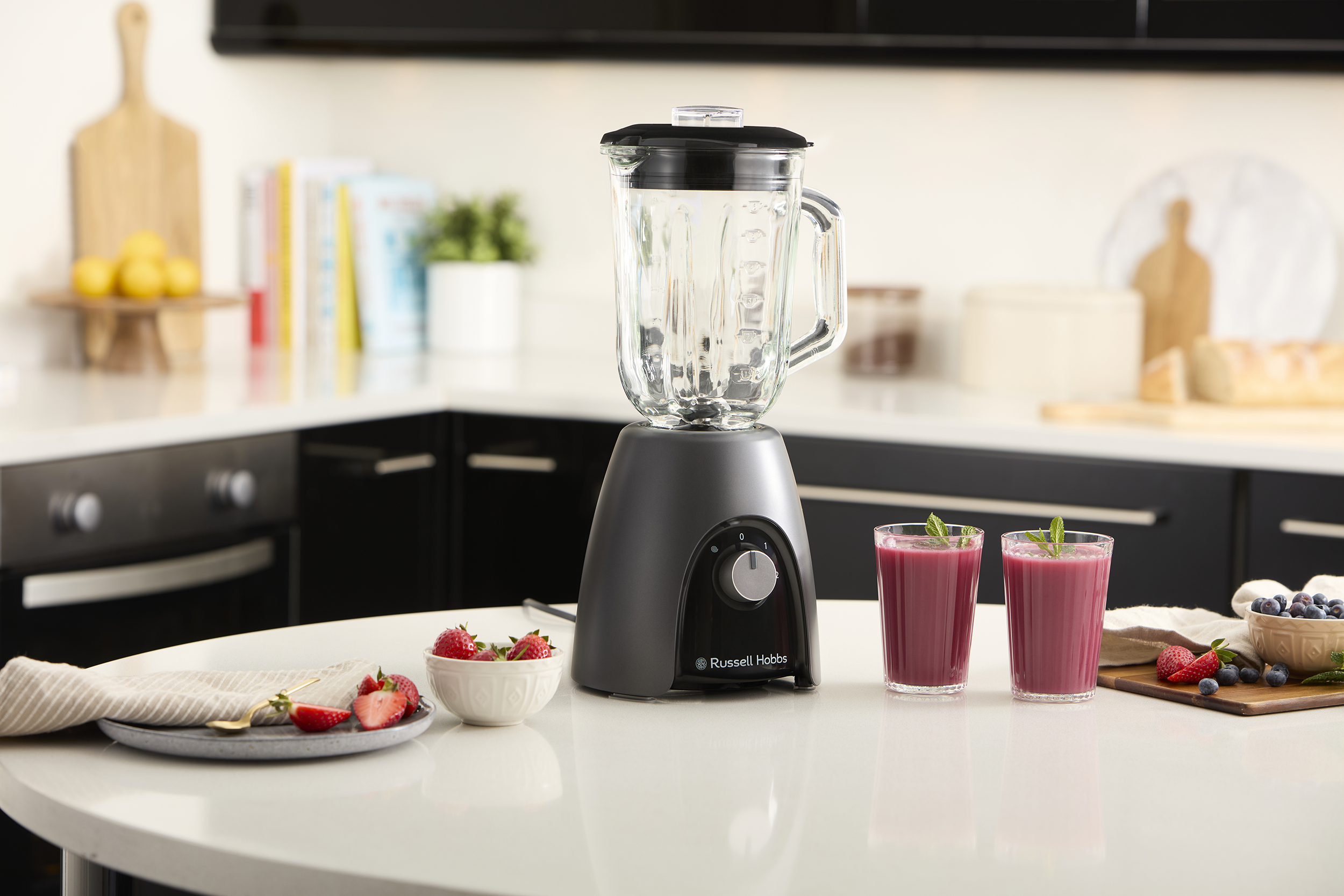 Szürke turmixgép üvegkancsóval, smoothie-kkal és gyümölcsökkel.