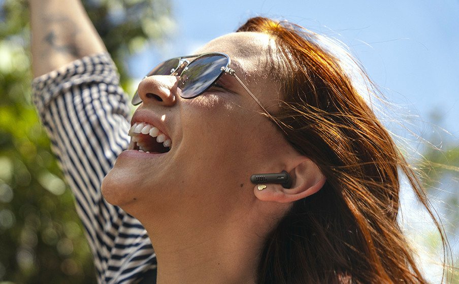 Femme avec lunettes de soleil sourit, écouteur dans l'oreille, bras levés, ciel bleu.
