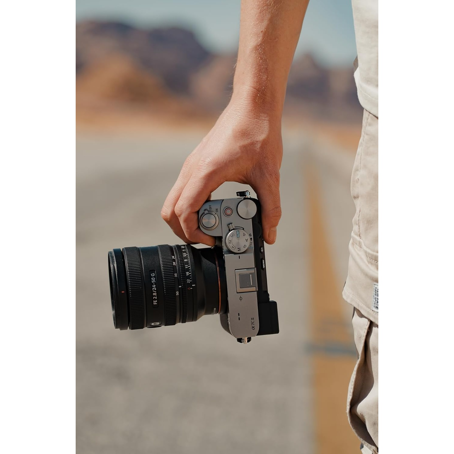 Una persona tiene in mano una fotocamera con obiettivo nero. Lo sfondo è sfocato con montagne.