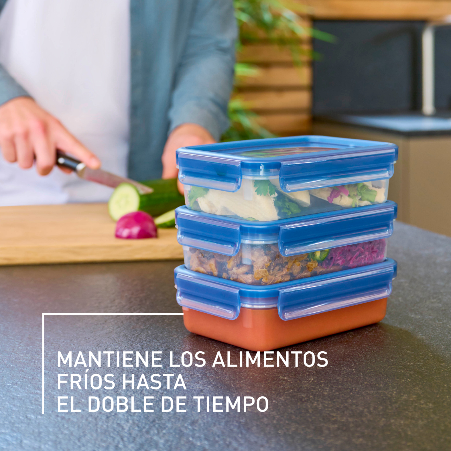 Tres recipientes de comida transparentes apilados con alimentos, una persona cortando verduras en el fondo.