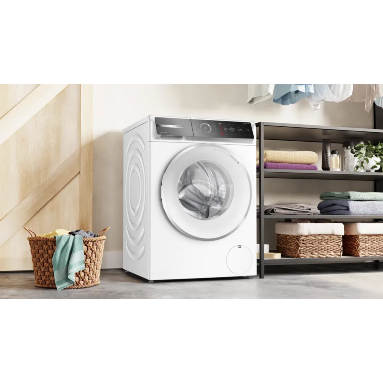 Een witte wasmachine in een keuken, met een mand met kleding en een plank met handdoeken in de buurt.