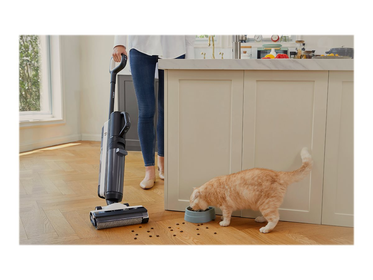Eine Frau saugt einen Holzboden. Eine orangefarbene Katze frisst aus einer Schüssel. Eine Kücheninsel ist sichtbar.
