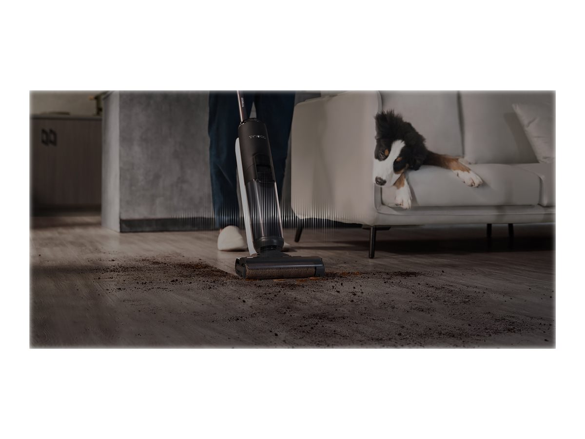 Eine Person saugt einen Holzboden; ein Hund ruht im Hintergrund auf einem Sofa.