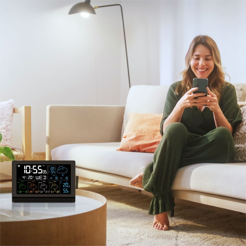 Kobieta siedzi na kanapie i korzysta z telefonu. Stacja pogodowa stoi na stole.