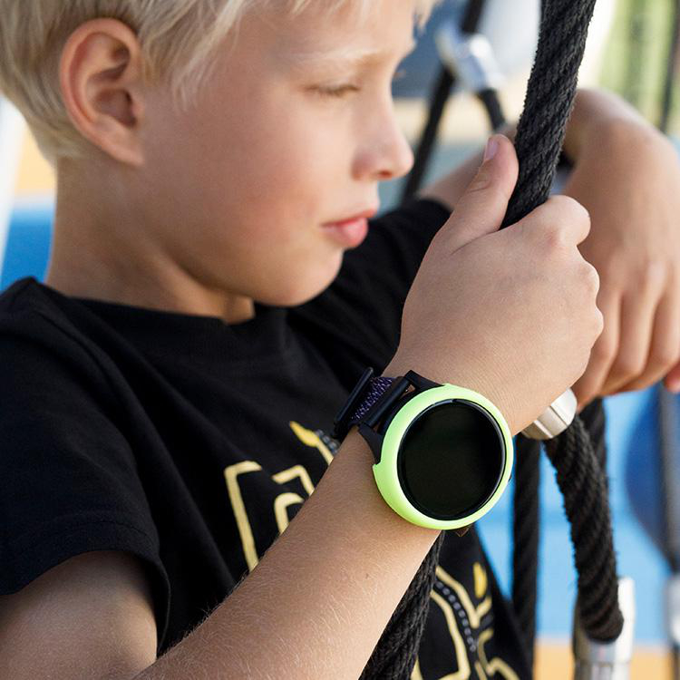 Blonder Junge mit Smartwatch mit grünem Rand, hält ein schwarzes Seil auf dem Spielplatz.