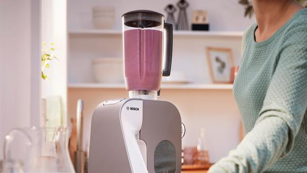 Mixer mit rosa Smoothie, Frau steht daneben in der Küche.
