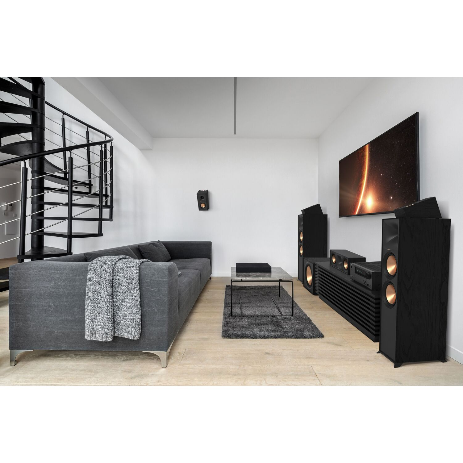 Woonkamer met luidsprekers, tv en grijze bank. Modern design met donker hout en een wenteltrap.