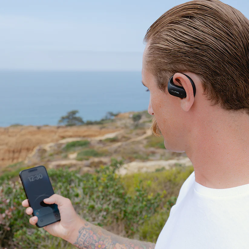 Ein Mann im Freien mit Ohrhörern benutzt ein Telefon. Der Hintergrund ist eine malerische Aussicht auf das Meer.