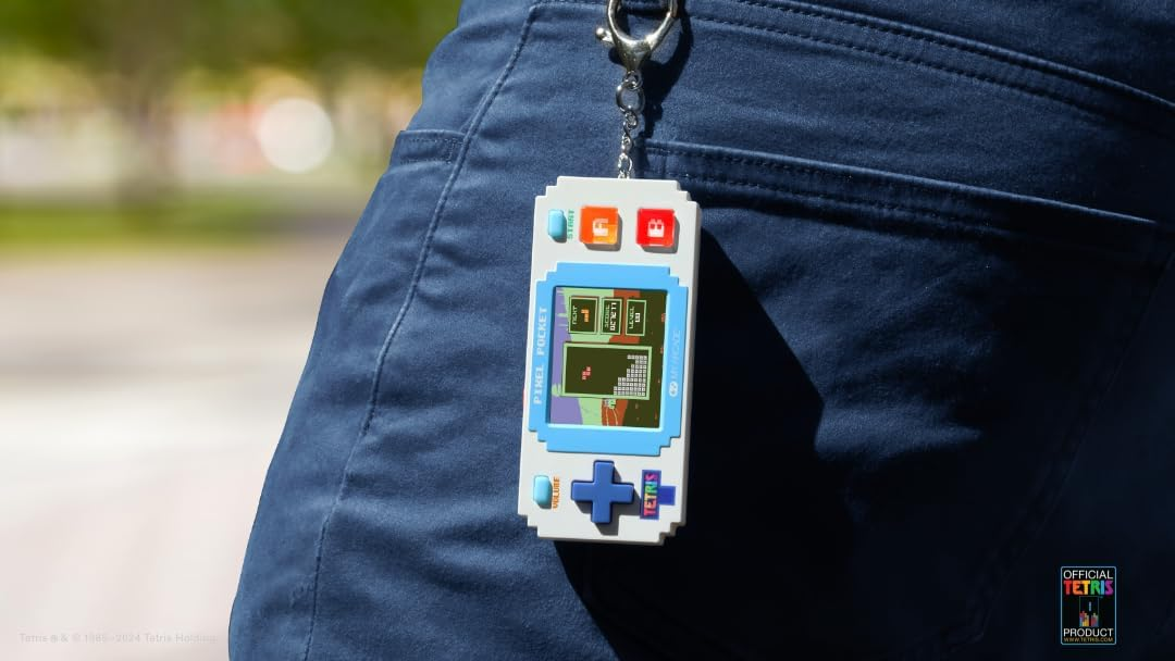 Un juego Pixel Pocket en un llavero, colgando de los jeans azules de una persona. El juego muestra Tetris.