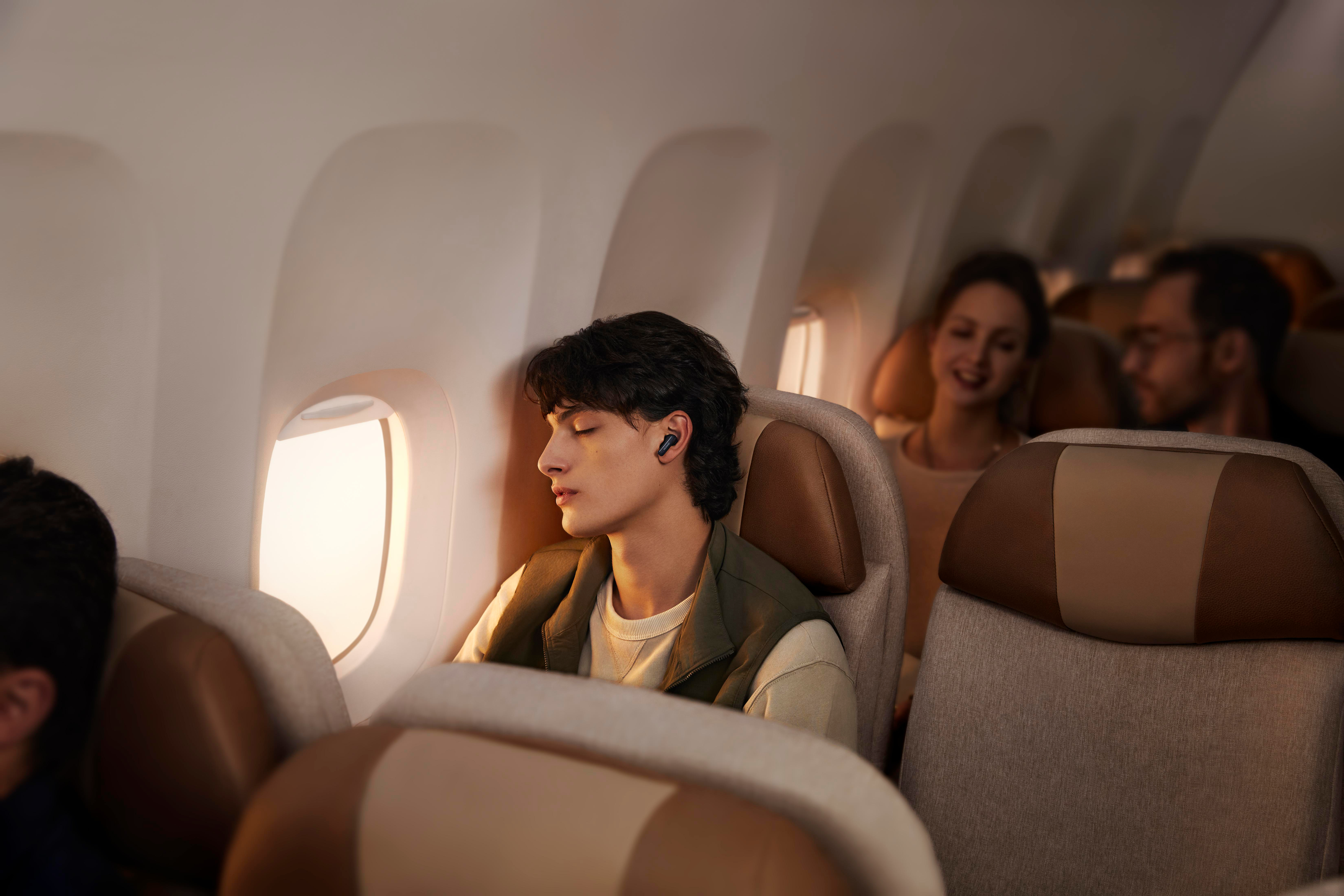 Mann mit Ohrhörern in einem Flugzeug, zwei weitere Passagiere im Hintergrund.