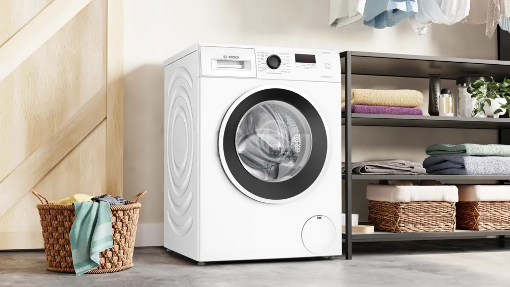 Biała pralka. Kosz na pranie znajduje się na pierwszym planie. Półka z przedmiotami po prawej.