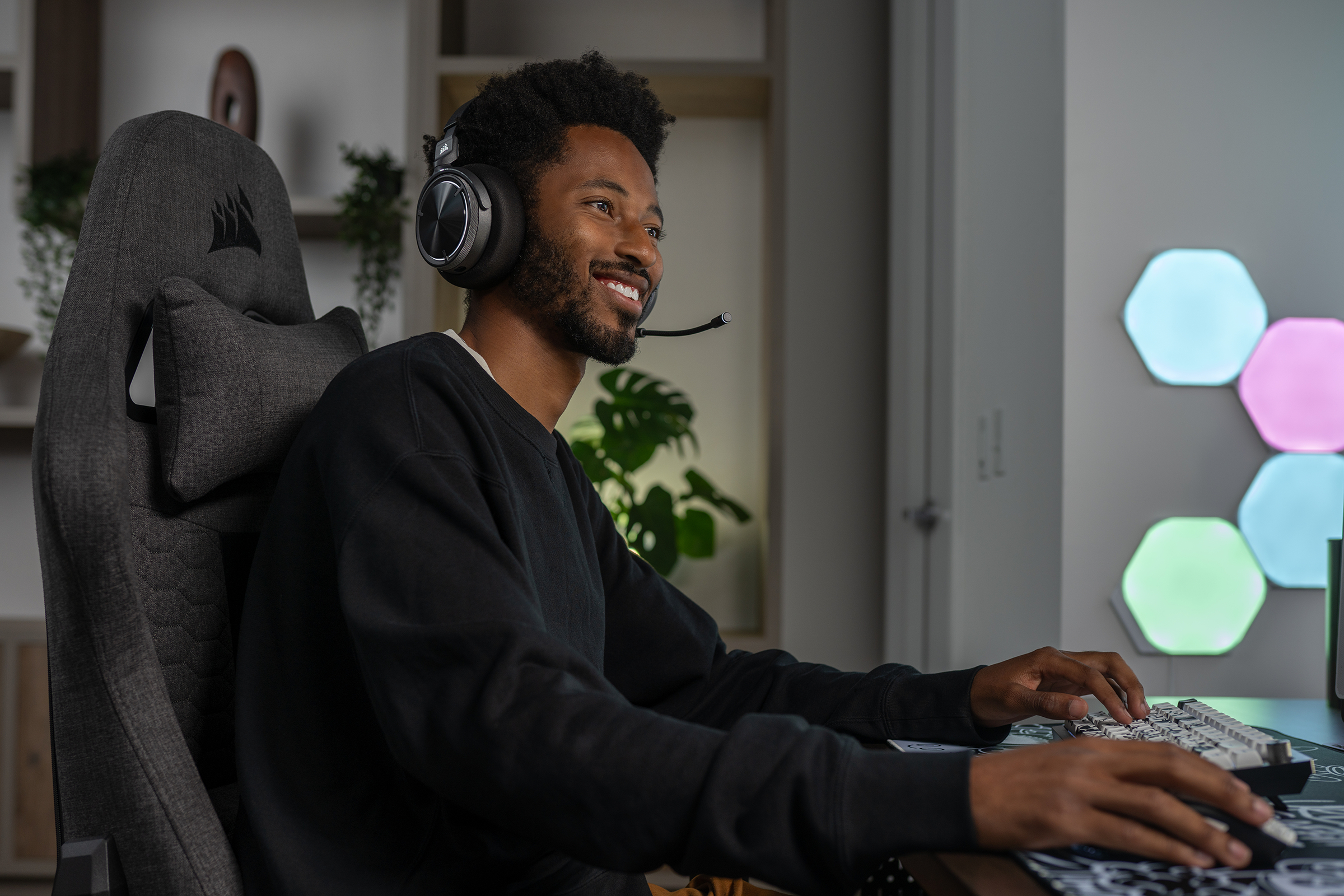 Ein Mann mit Headset lächelt beim Spielen am Computer. Der Raum hat einen Gaming-Stuhl und bunte Hexagon-Lichter.