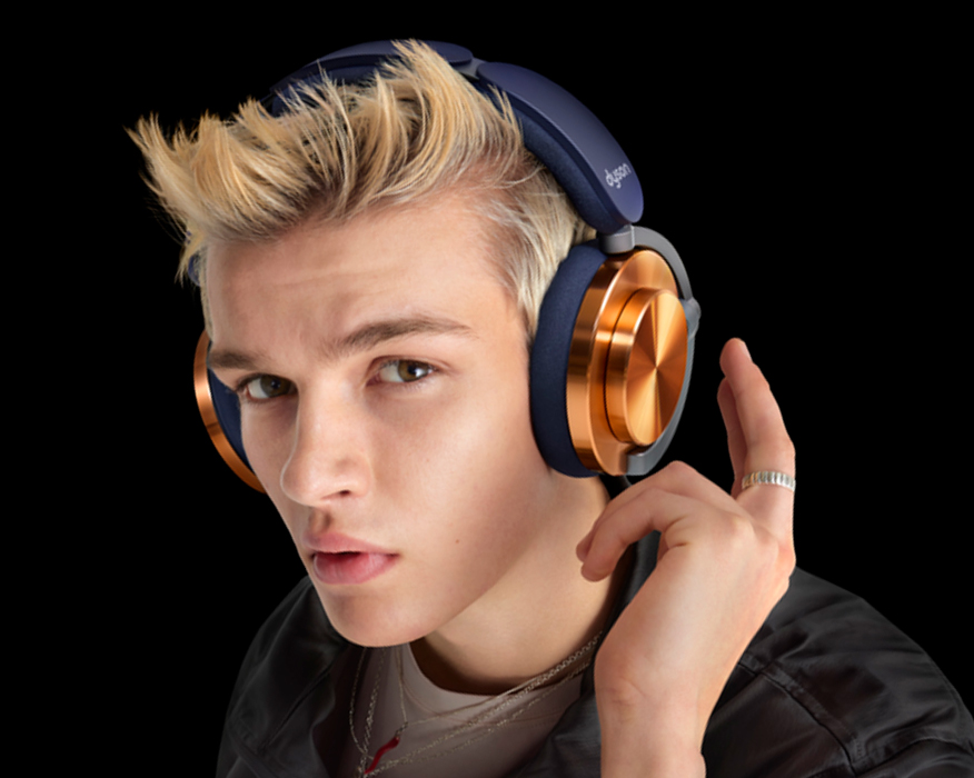 Persona rubia con auriculares. Los auriculares son azul marino y dorados. Fondo negro.