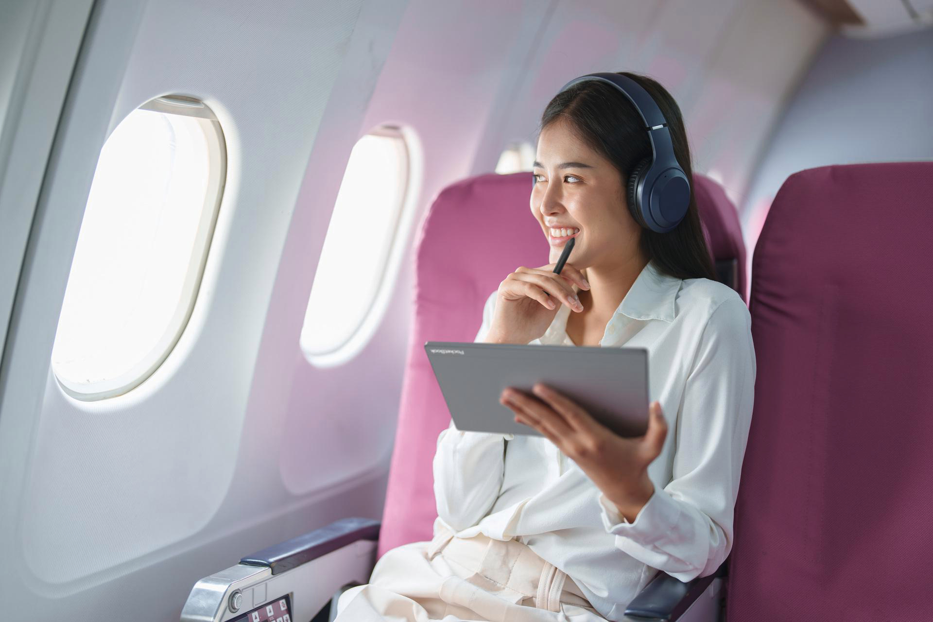 Eine Frau mit Kopfhörern in einem Flugzeug, die ein Tablet benutzt und am Fenster lächelt.