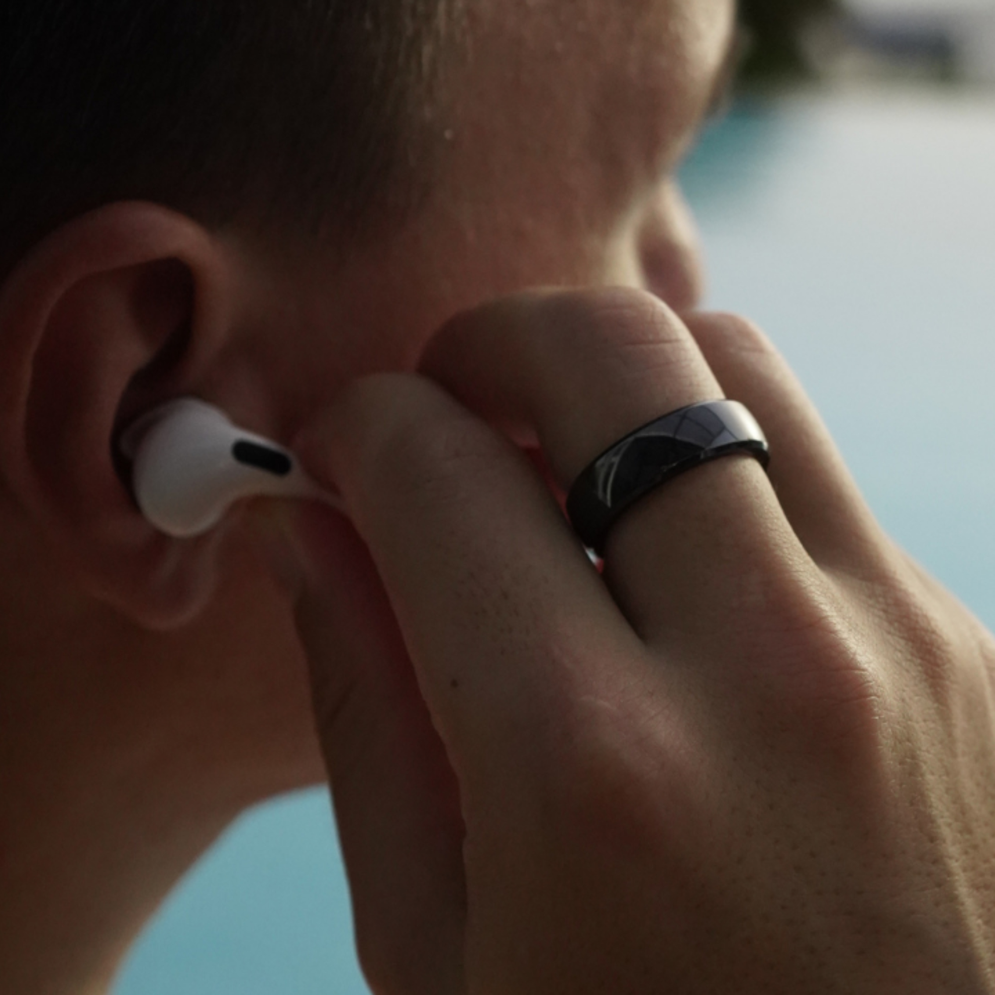 Eine Hand mit schwarzem Ring, die einen Ohrhörer einsetzt, mit verschwommenem Pool im Hintergrund.