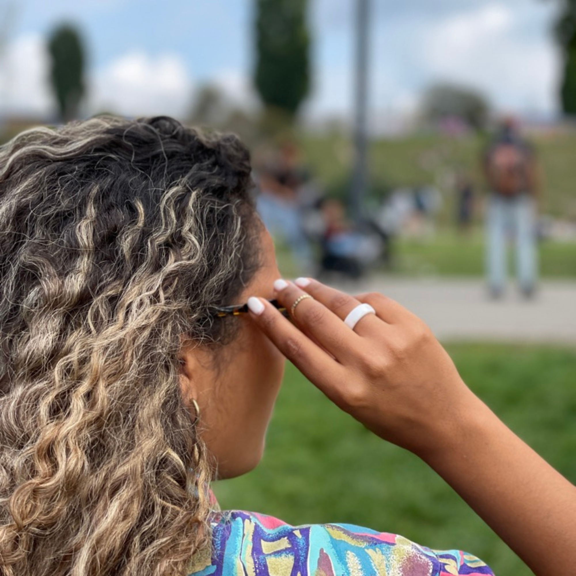 Eine Frau mit lockigem Haar berührt ihr Ohr. Sie trägt Ring und Ohrringe. Personen im Hintergrund.