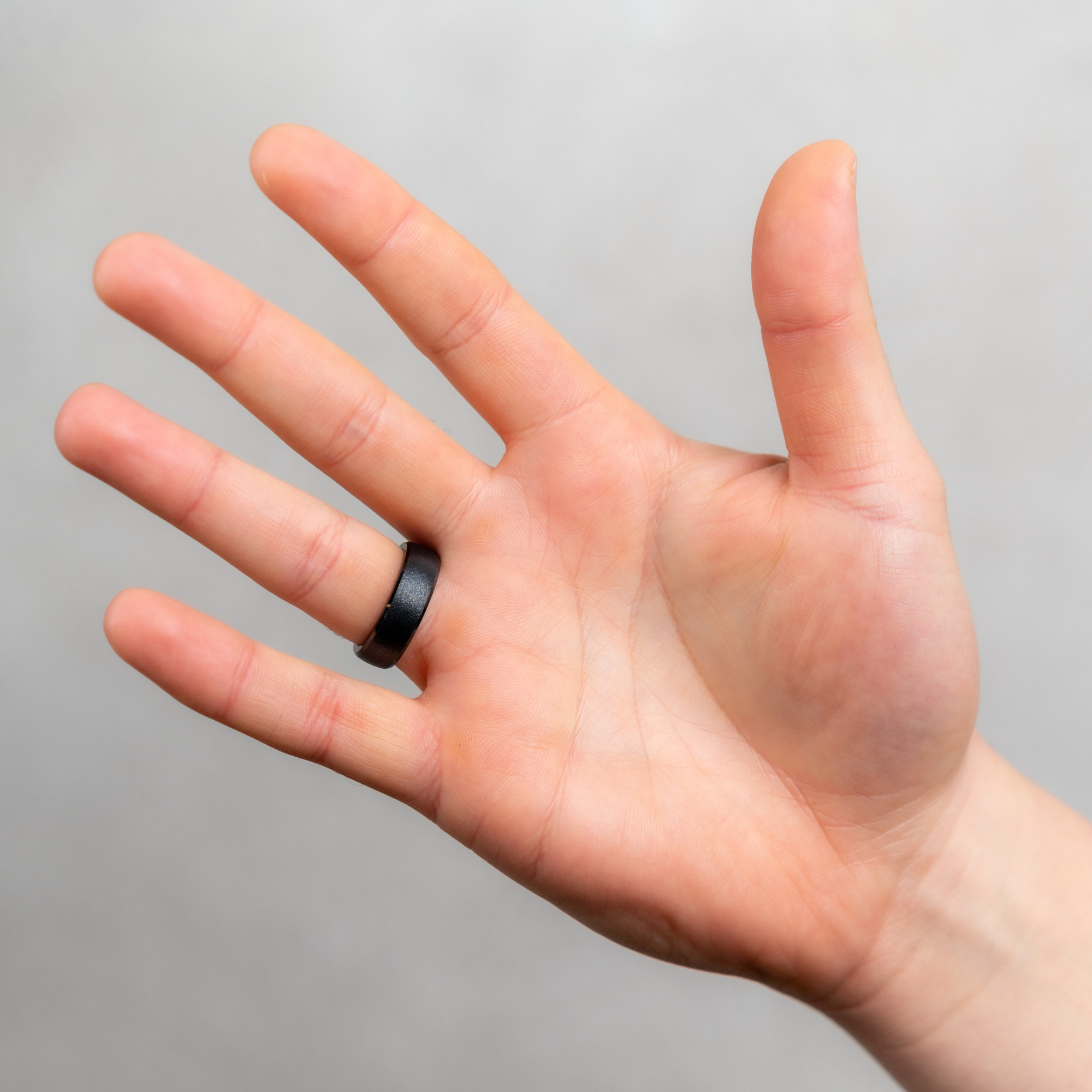 Eine Hand, die einen schwarzen Ring am Ringfinger trägt. Die Hand ist vor grauem Hintergrund geöffnet.