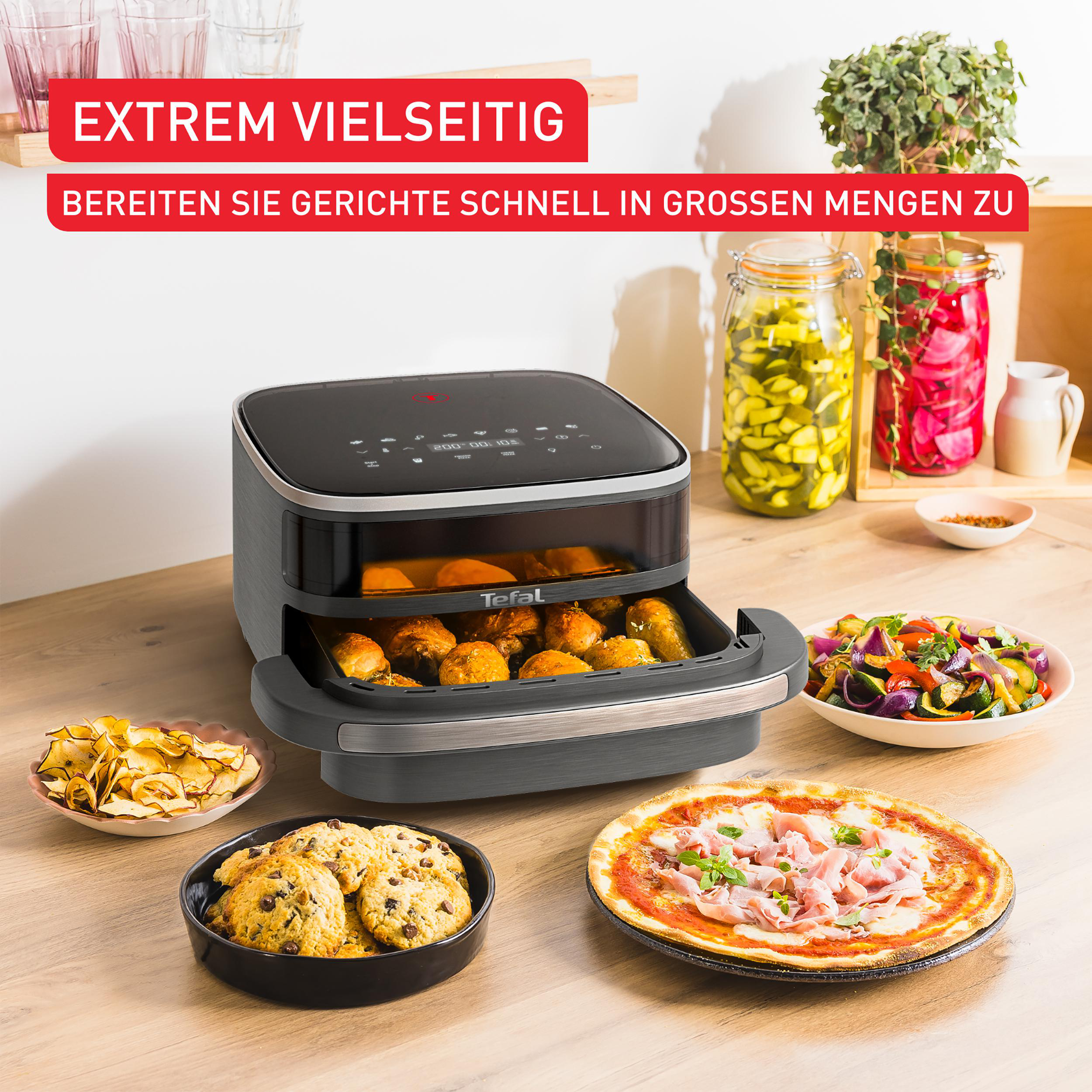 Heißluftfritteuse mit Essen. Snacks und eine Pizza auf einem Tisch. Weitere Lebensmittel im Hintergrund.