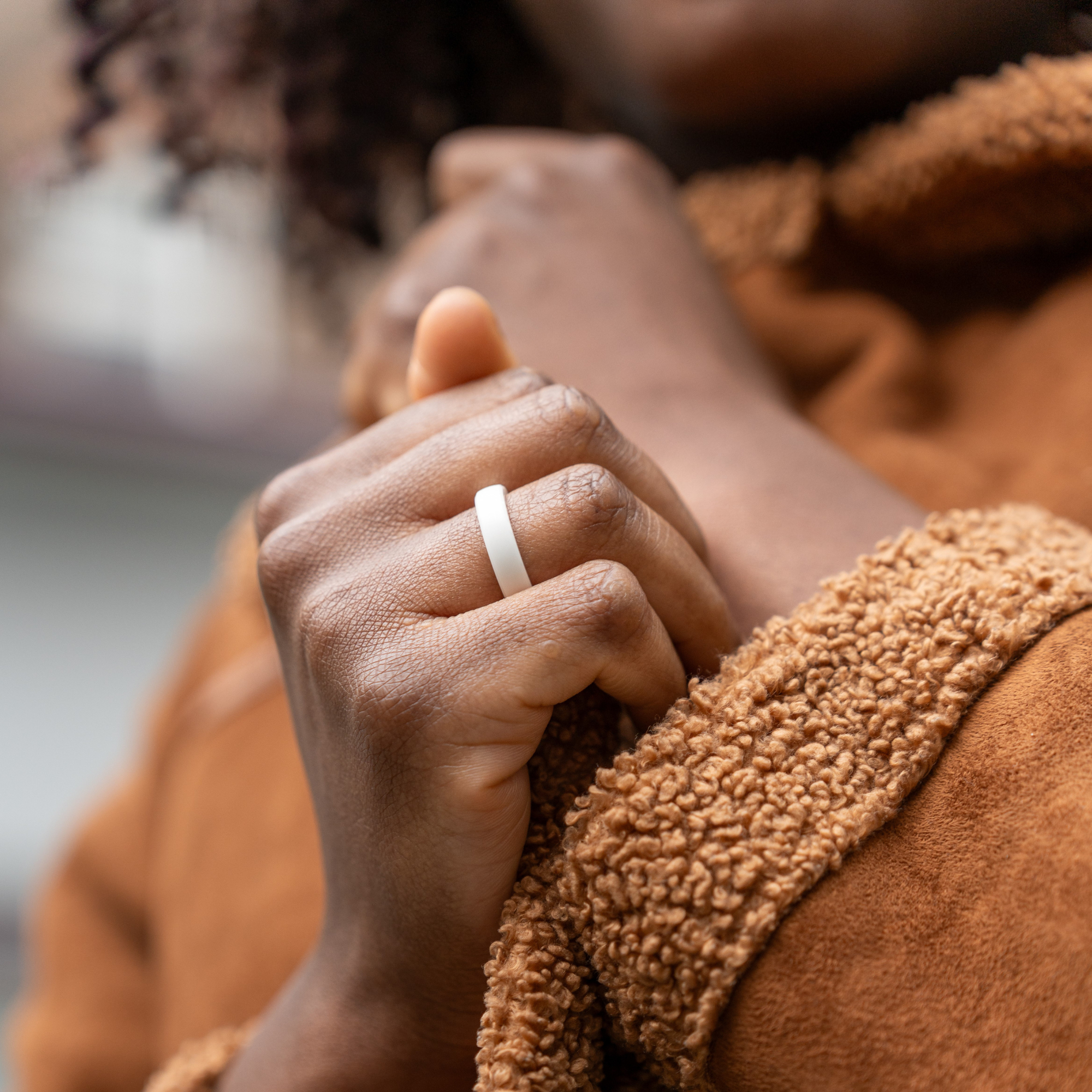 Eine Person trägt einen weißen Ring am Finger. Sie trägt einen braunen Mantel.