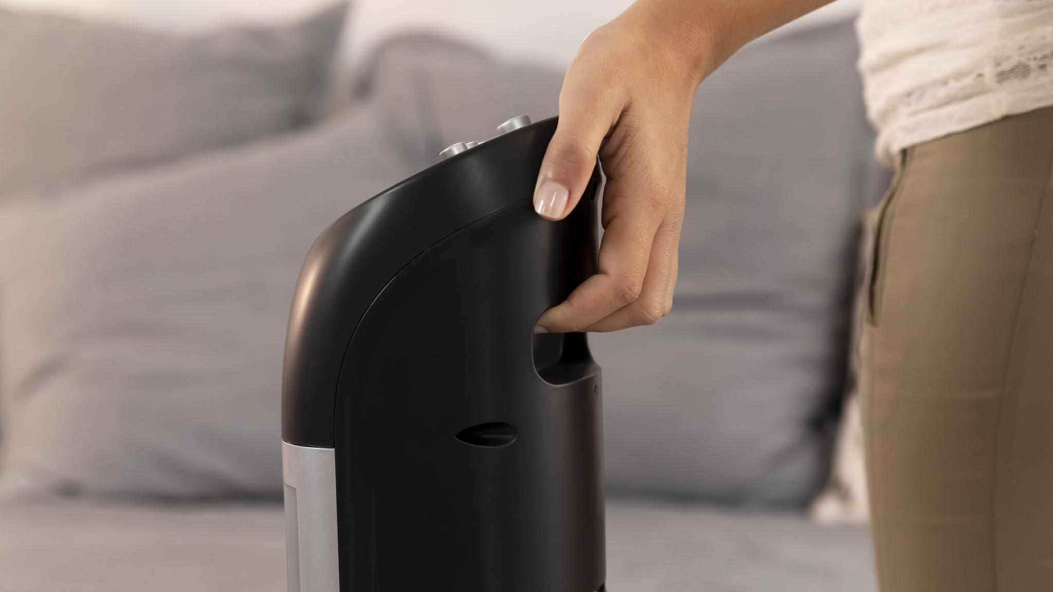 Una mano sosteniendo un purificador de aire negro, con un sofá gris borroso al fondo.
