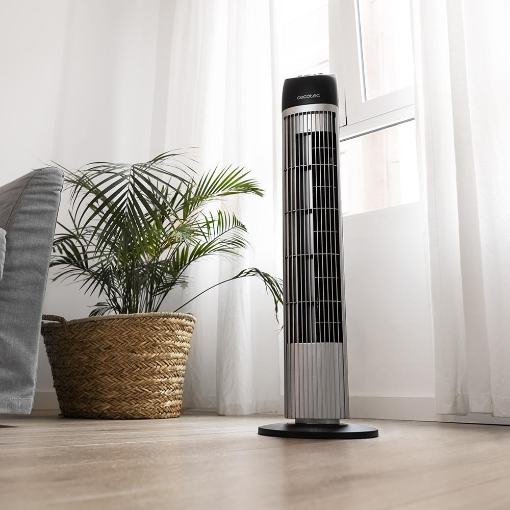 Ventilador alto plateado y negro junto a una cesta con una planta. El ventilador está frente a una ventana.