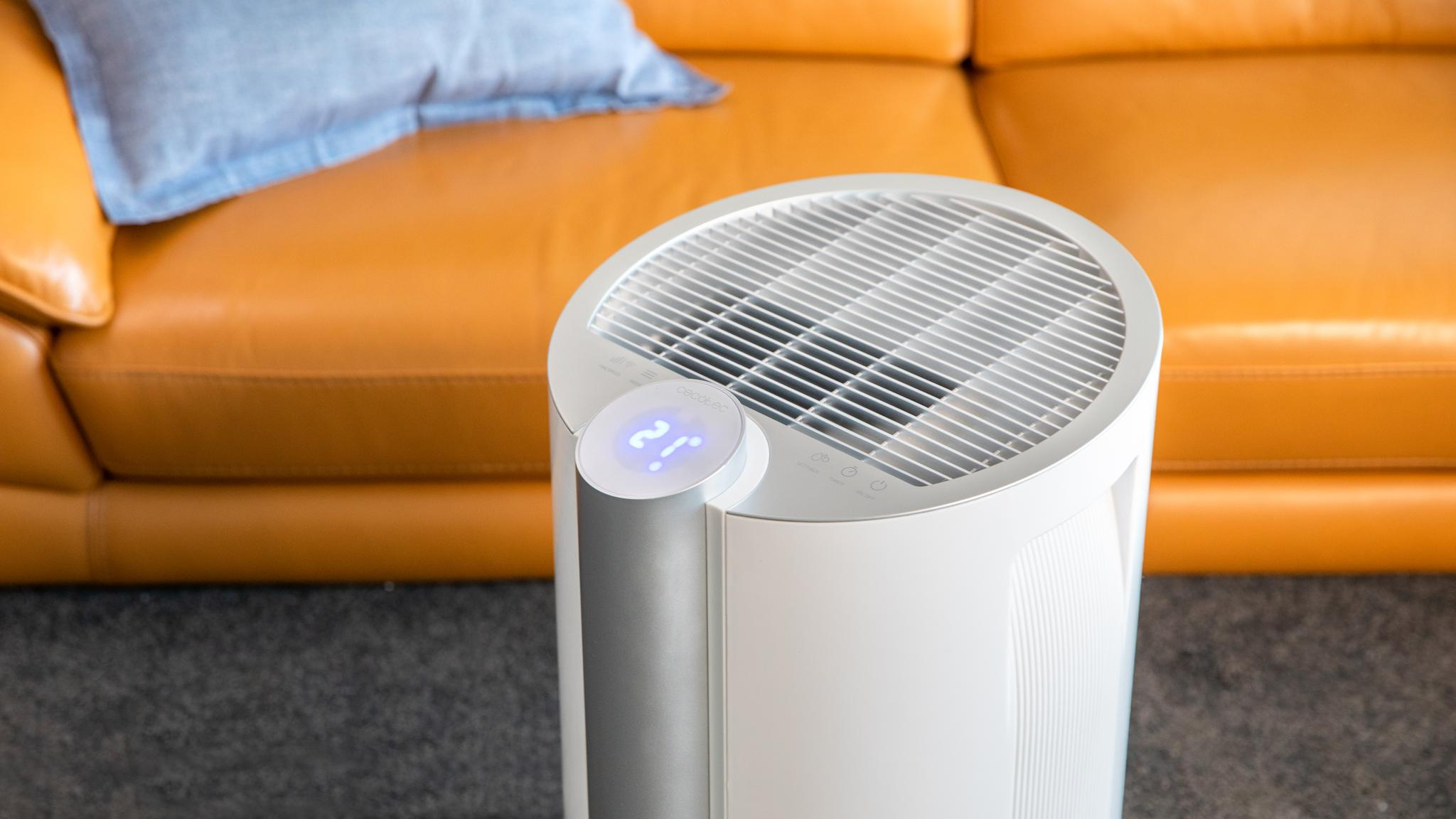 Purificador de aire blanco con pantalla digital, colocado en una habitación con sofá de cuero y suelo alfombrado.