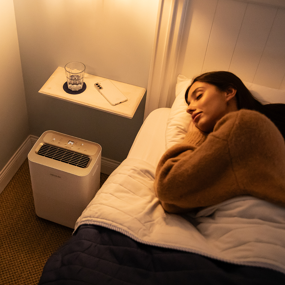 Una mujer durmiendo en una cama junto a un purificador de aire blanco.
