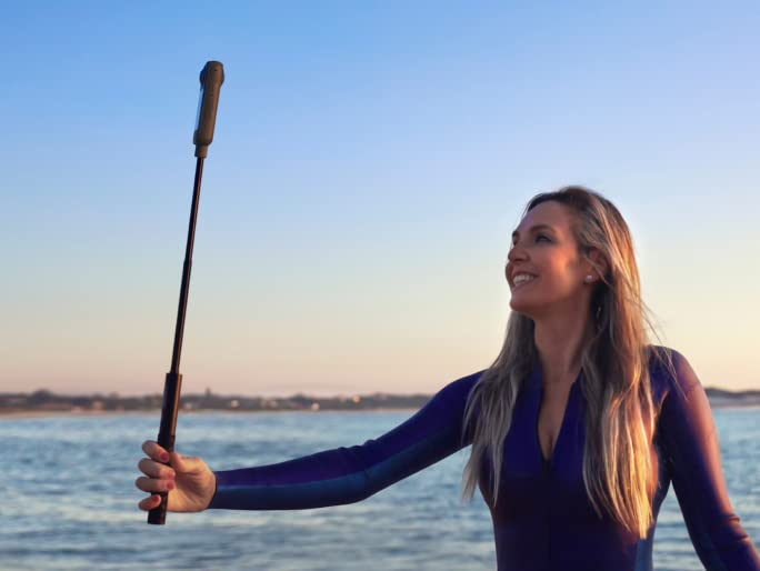 Mujer sosteniendo un palo selfie con cámara en la playa. Ella está sonriendo.