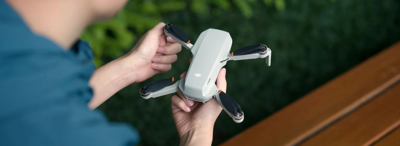 Osoba trzymająca szaro-czarny dron ze złożonymi ramionami, przed drewnianą ławką.