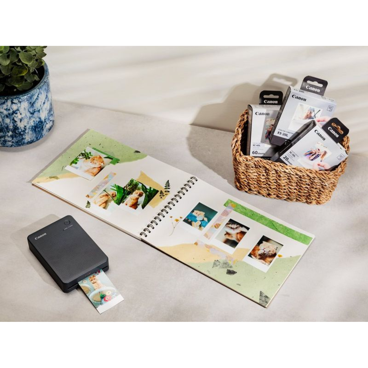 Photo printer, photos in a notebook, ink cartridges in a basket, all on a light surface.