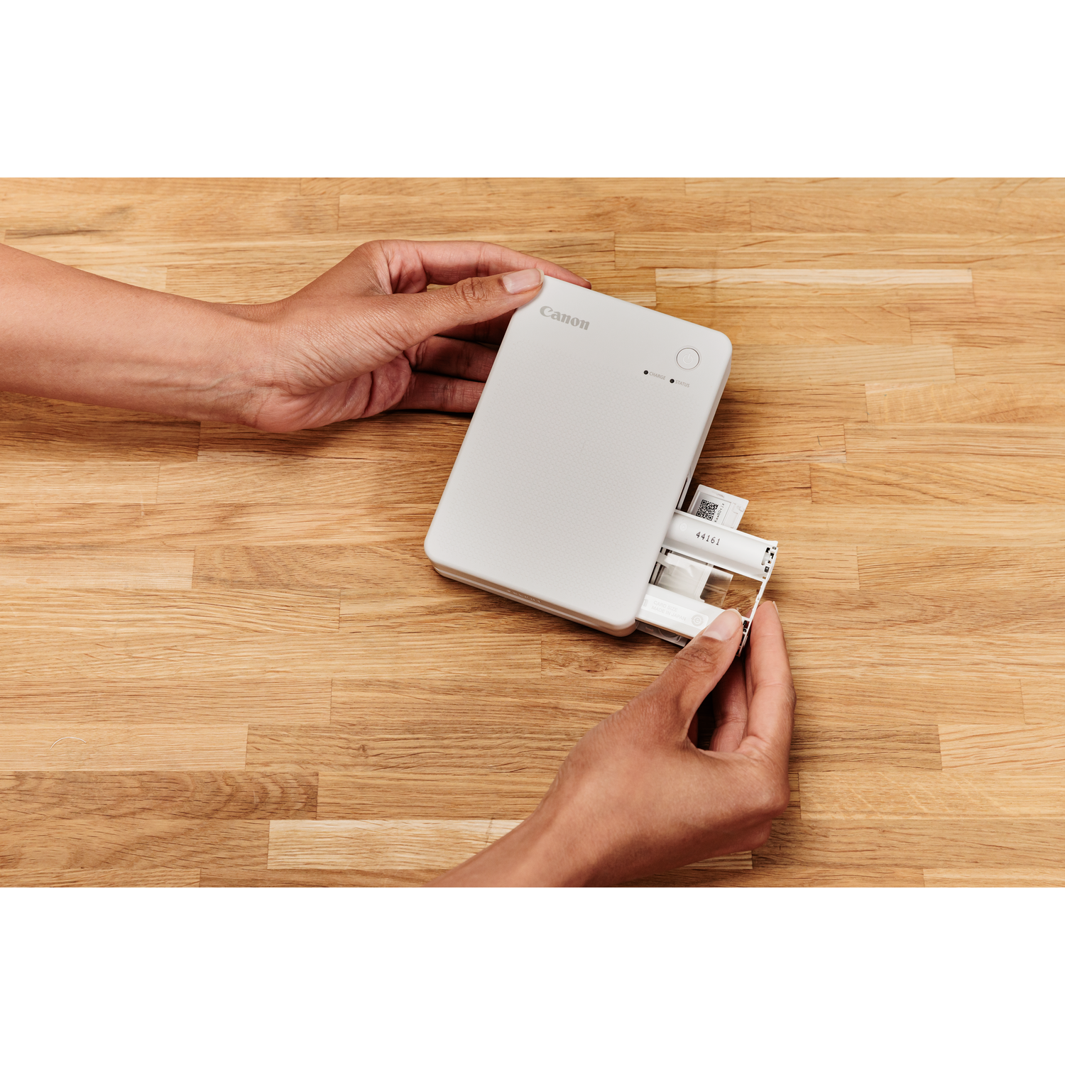 Hands inserting photo paper into a white Canon printer on a wooden table.