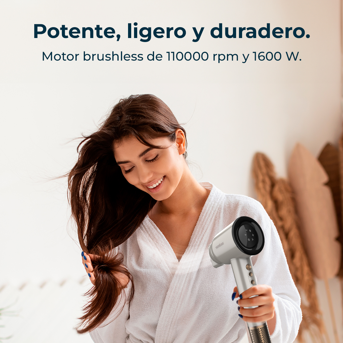 Mujer usando secador plateado, sonriendo. Fondo: pared blanca y plantas secas.