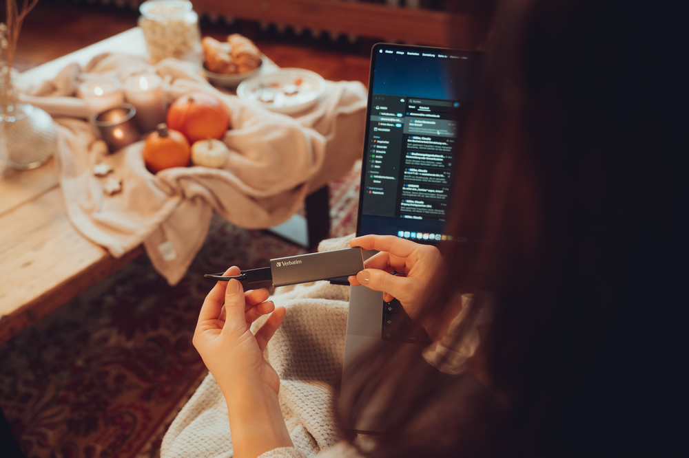 Egy nő egy szürke Verbatim eszközt tart, ami egy laptophoz van csatlakoztatva, hangulatos háttérrel.