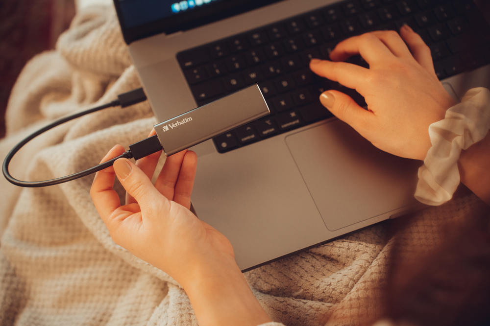 Kezek egy ezüst USB meghajtót tartanak, laptopba dugva, takarón.
