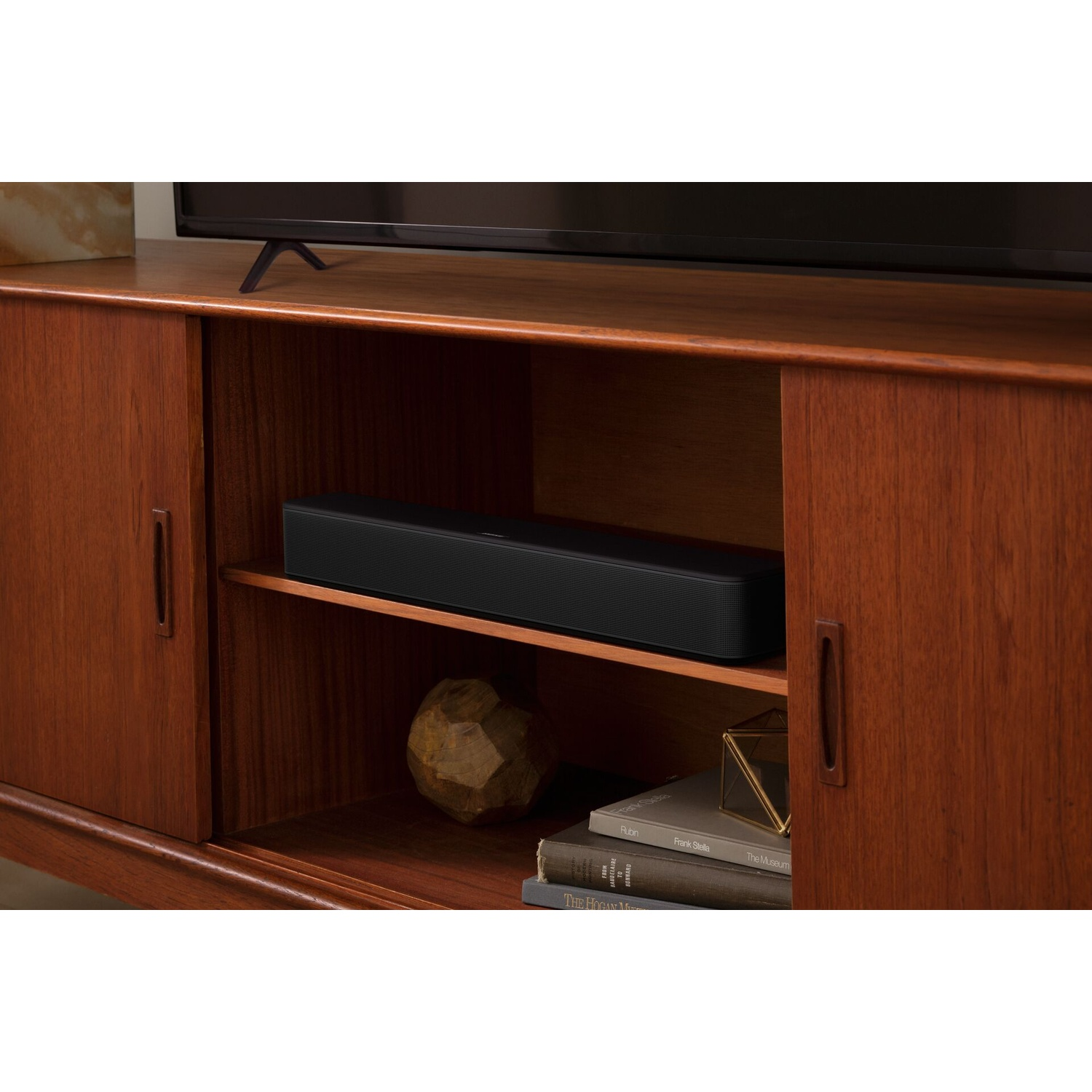 Una soundbar nera è su uno scaffale in un mobile di legno. Libri e una pietra decorativa vicini.