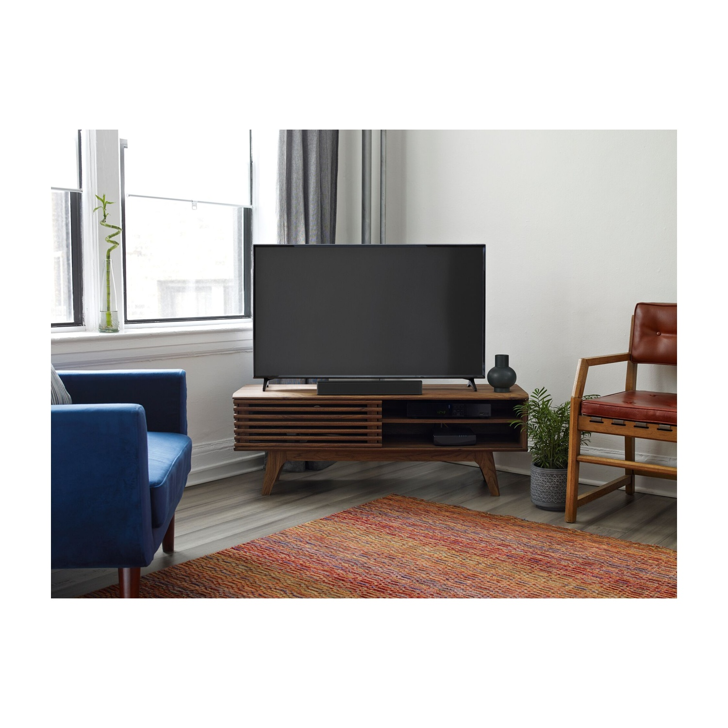 Soggiorno con una TV su un mobile in legno. Una poltrona blu e una sedia marrone. Un tappeto sul pavimento. Finestra a sinistra.