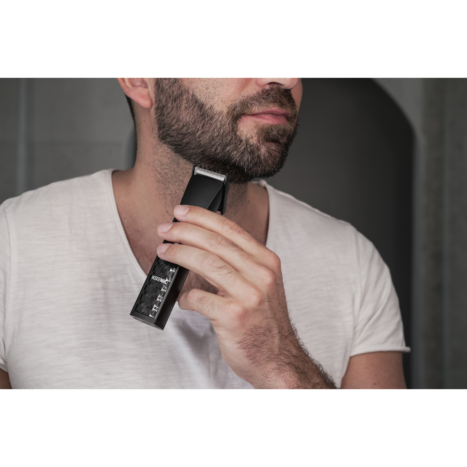 Man trimming beard with black beard trimmer. White shirt, stubble beard, neutral background.