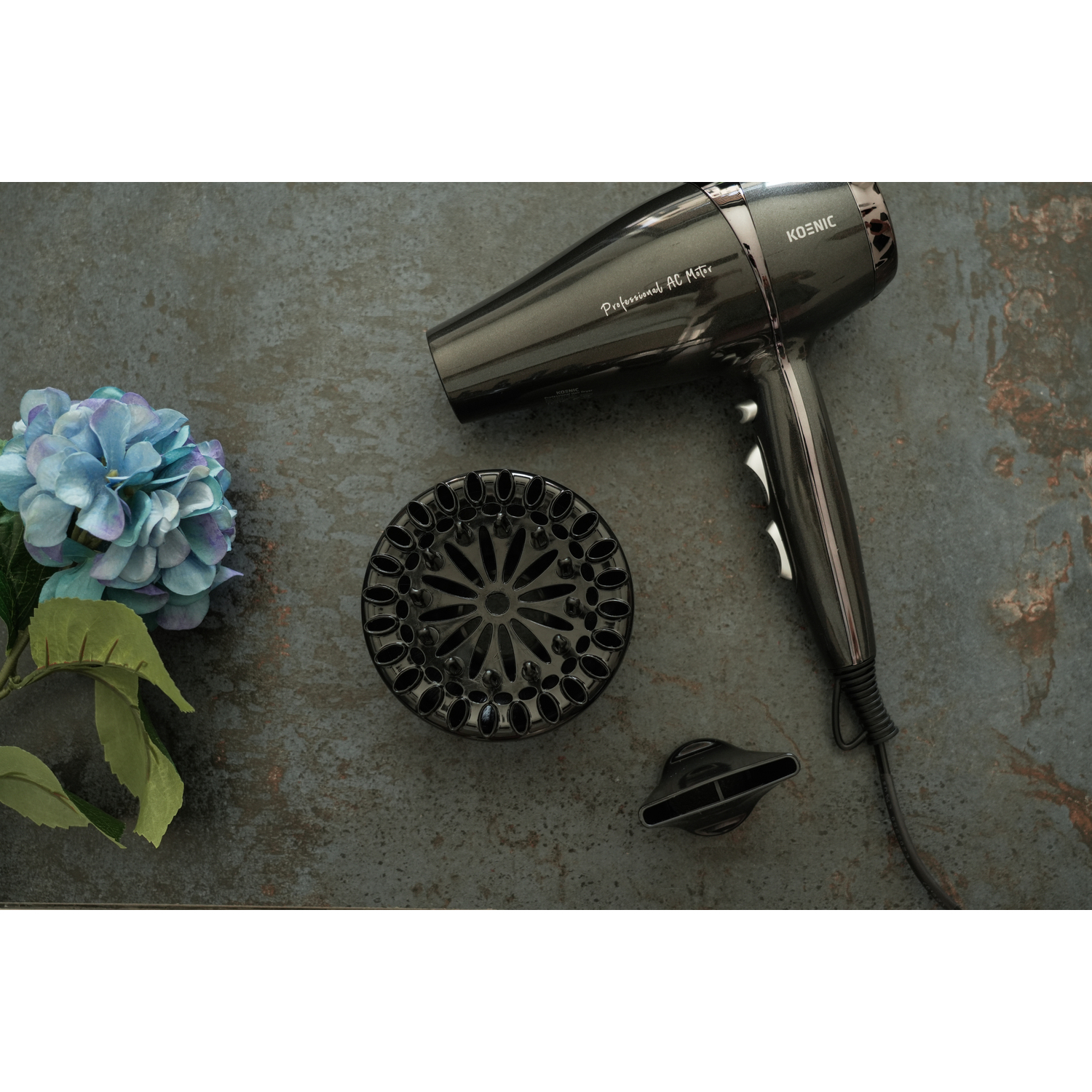 Black hair dryer with attachments, beside a blue flower on a textured grey surface.