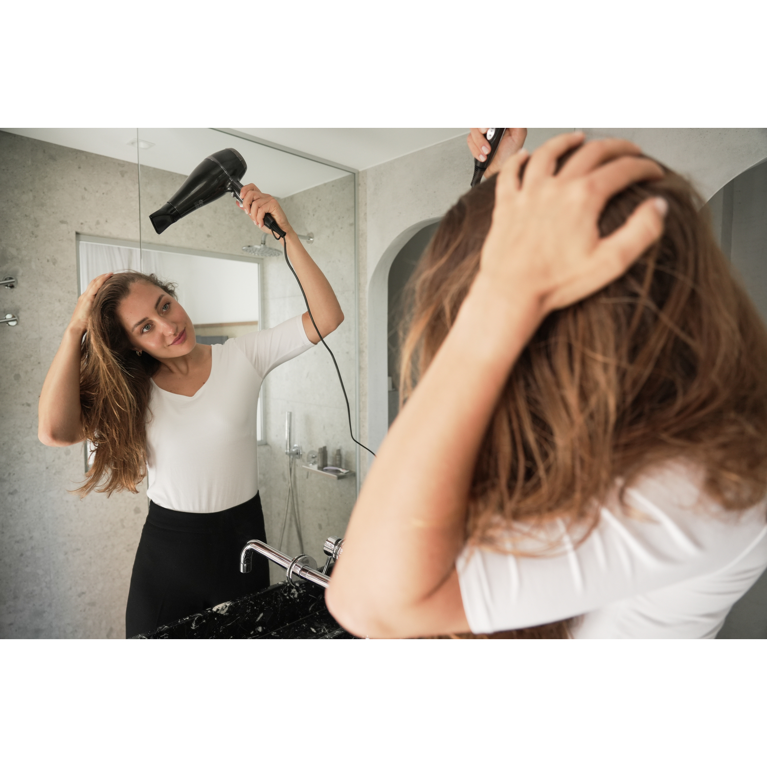 A woman drying her hair in front of a mirror. She wears a white shirt and black skirt. Bathroom setting.