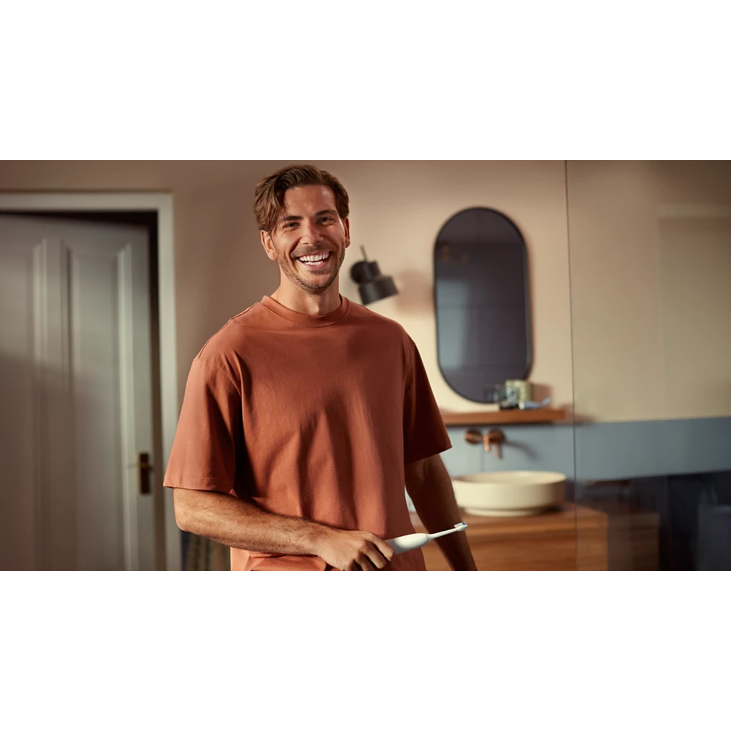 Un homme souriant, tenant une brosse à dents électrique blanche dans une salle de bain.