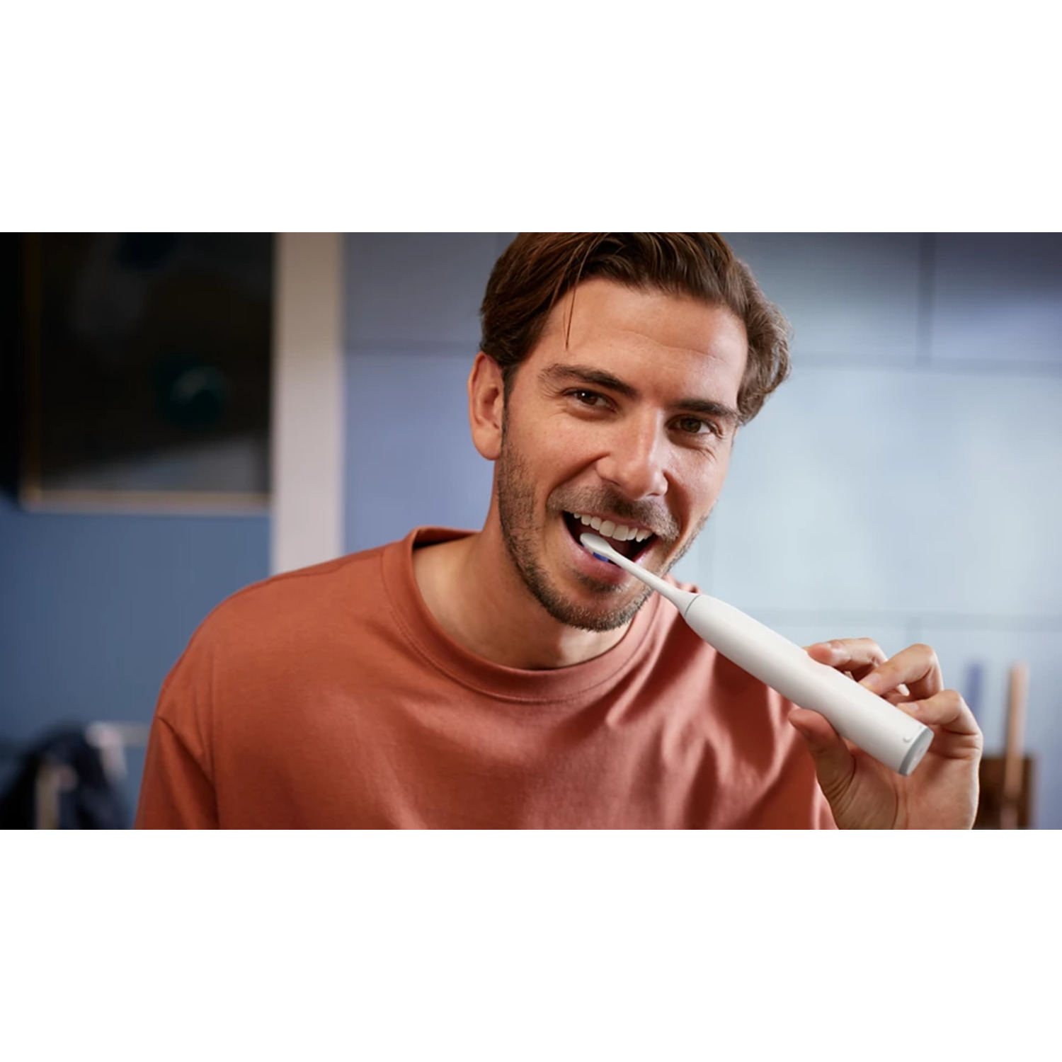 Homme se brossant les dents avec une brosse à dents électrique blanche. Souriant, dans salle de bain. Chemise marron.