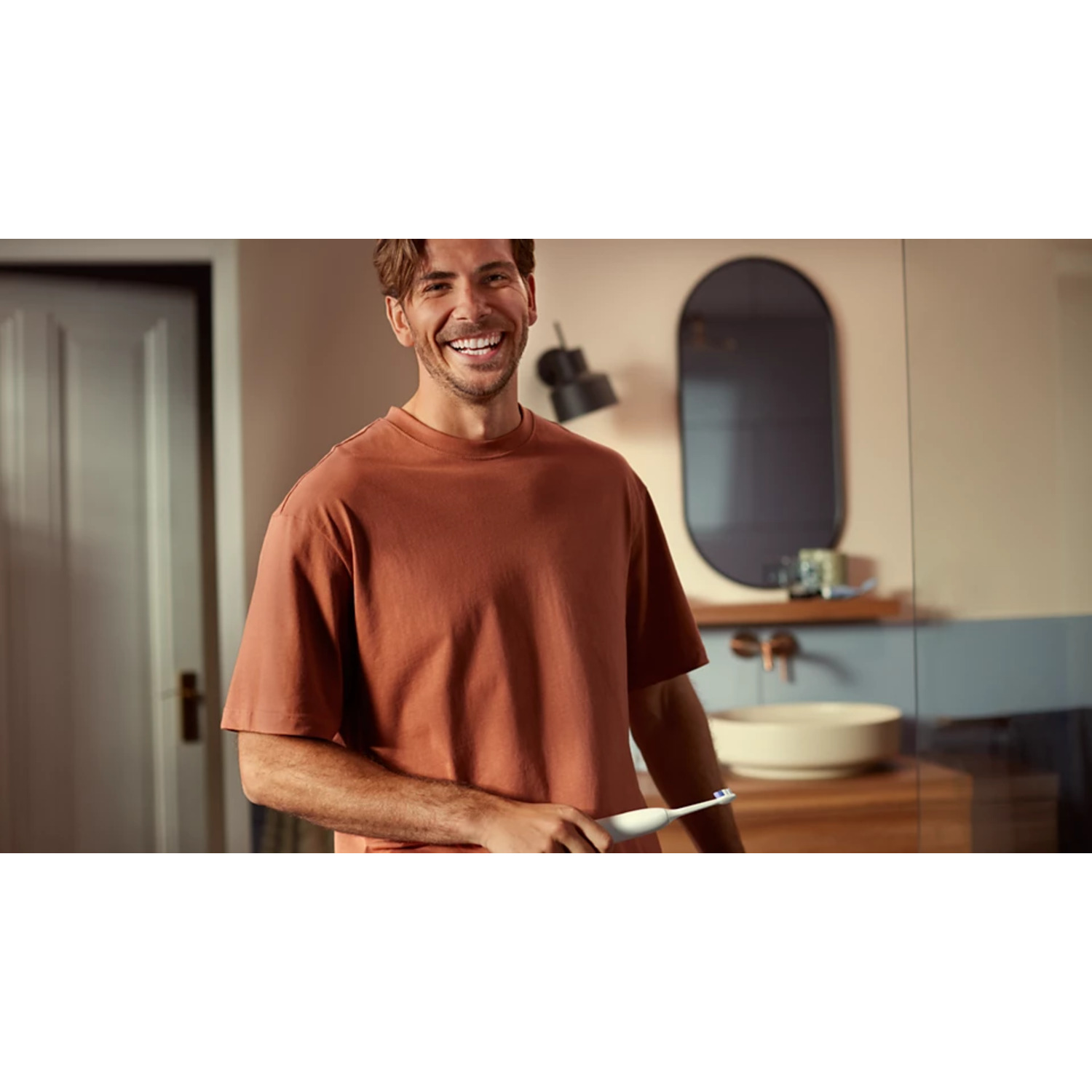 Un homme souriant, tenant une brosse à dents électrique blanche dans une salle de bain.
